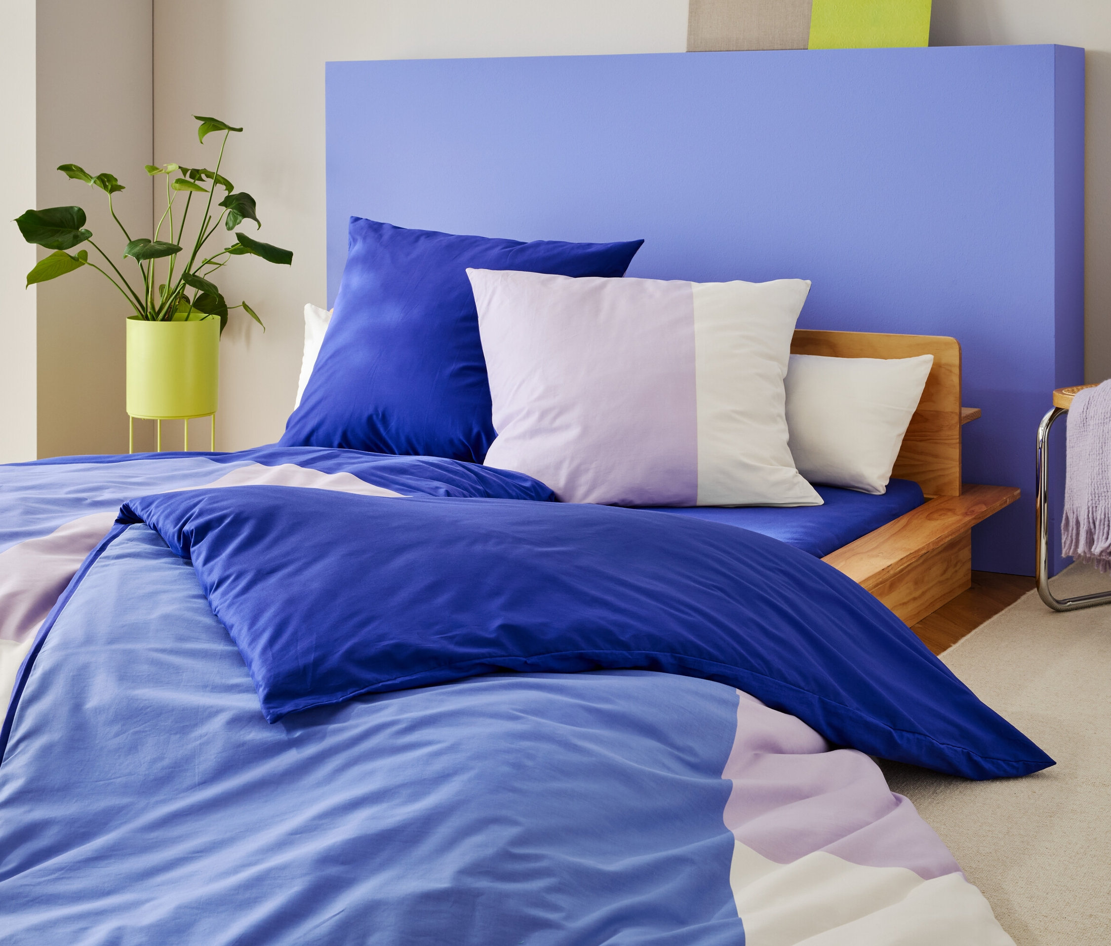 Posteľ s modro-fialovou posteľnou bielizňou a vankúšmi stojí pred modrou stenou, vedľa nej rastlina a stolička s dekou.