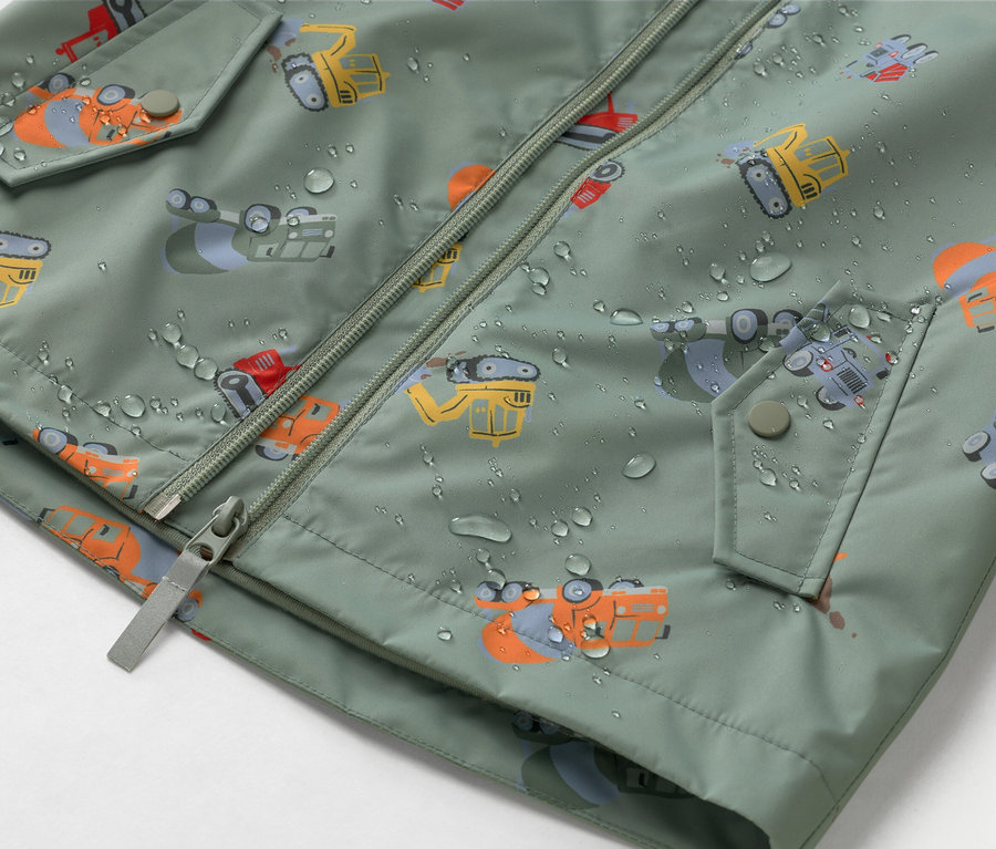 Detailný záber olivovozelenej bundy so zipsom a motívom stavebných strojov, pokrytej kvapkami vody.