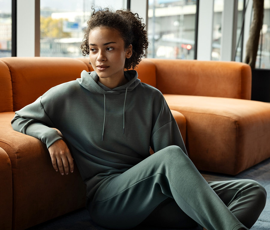 Žena sedí na zemi, opiera sa o oranžovú sedačku a má na sebe zelenú mikinu s kapucňou a tepláky.