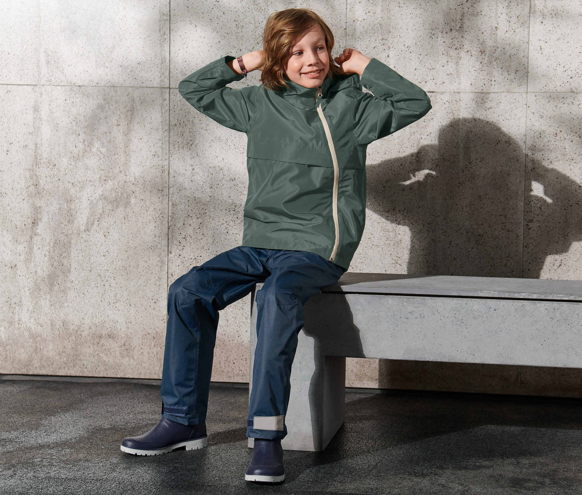 Chlapec sedí na lavičke a upravuje si kapucňu bundy do každého počasia.