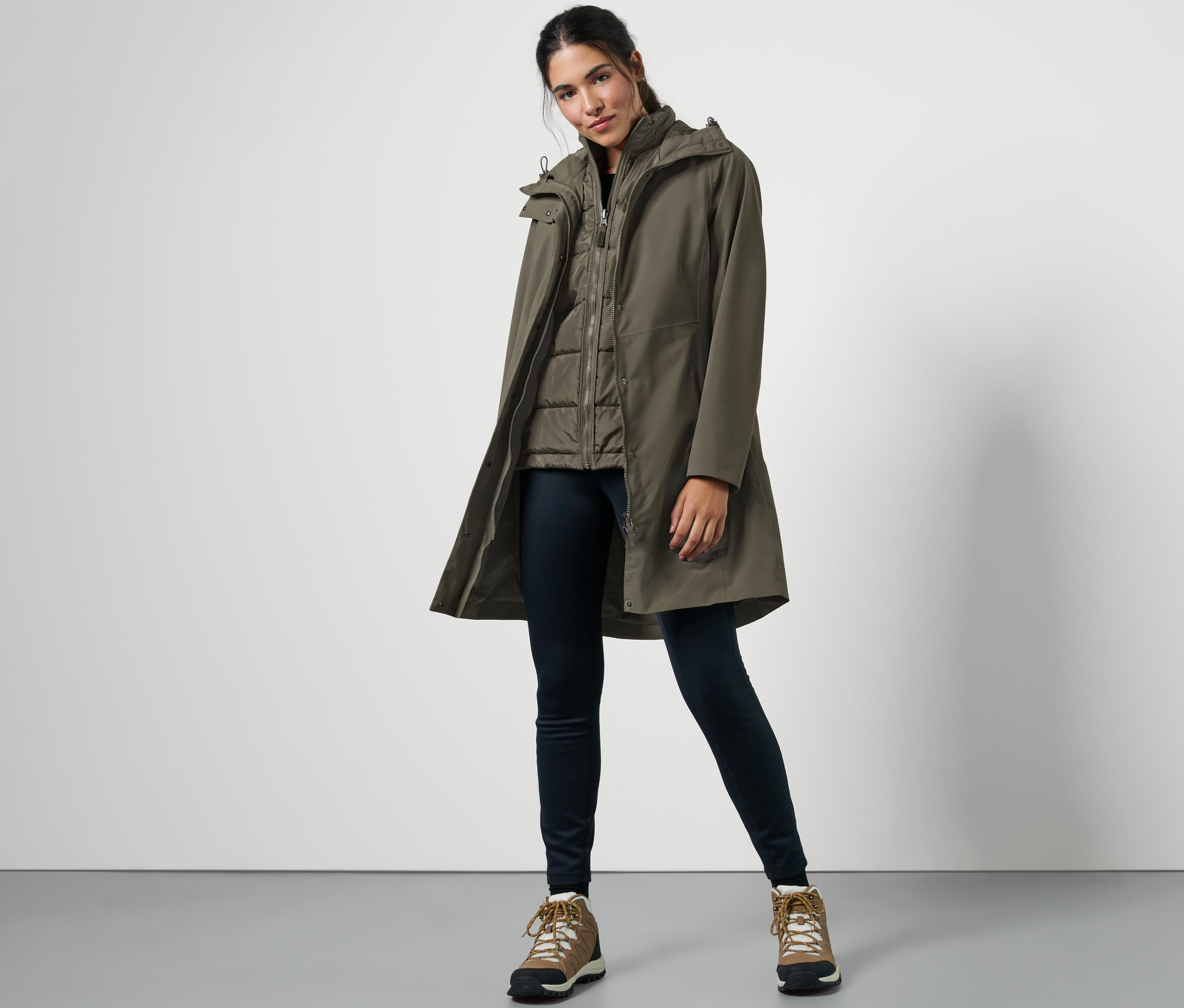 Žena pózuje v kabáte do dažďa s vestou 3 v 1, tmavých džínsoch a turistických topánkach.