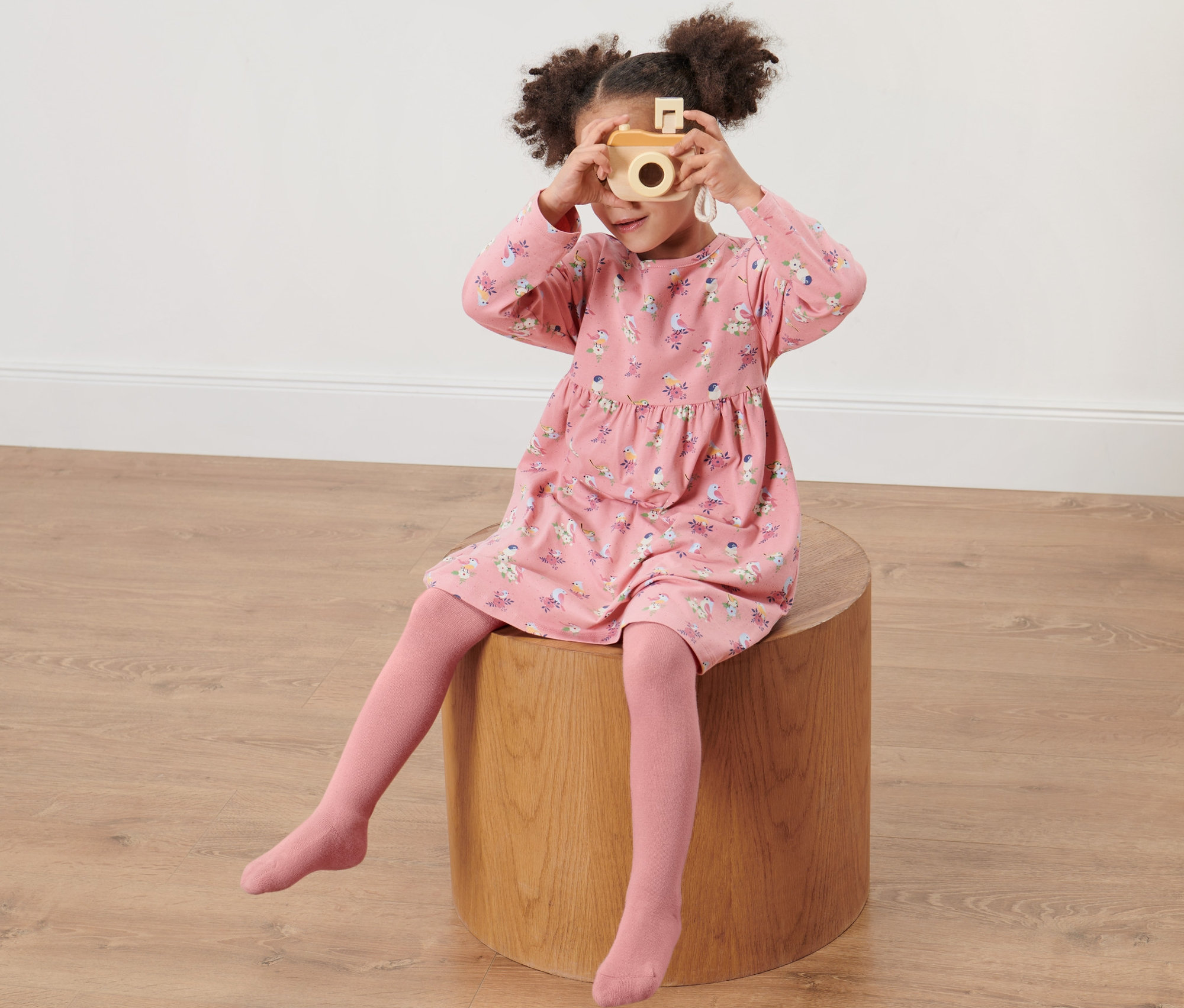 Dievča sedí na drevenej stoličke a fotí dreveným fotoaparátom. Má na sebe ružové šaty s potlačou a detské pančuchy, 2 ks, ružové/svetlomodré.