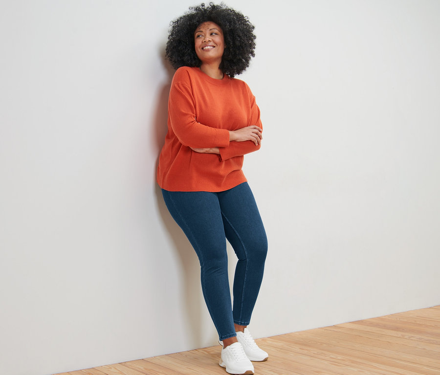 Žena sa opiera o stenu a má na sebe pulóver z jemnej pleteniny s kašmírom, džínsy a tenisky.