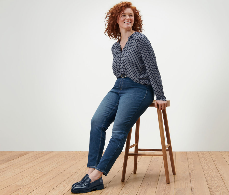 Žena sedí na drevenej stoličke a má na sebe blúzkové tričko s potlačou, tregínsy »Fit Nelly« a kožený opasok.