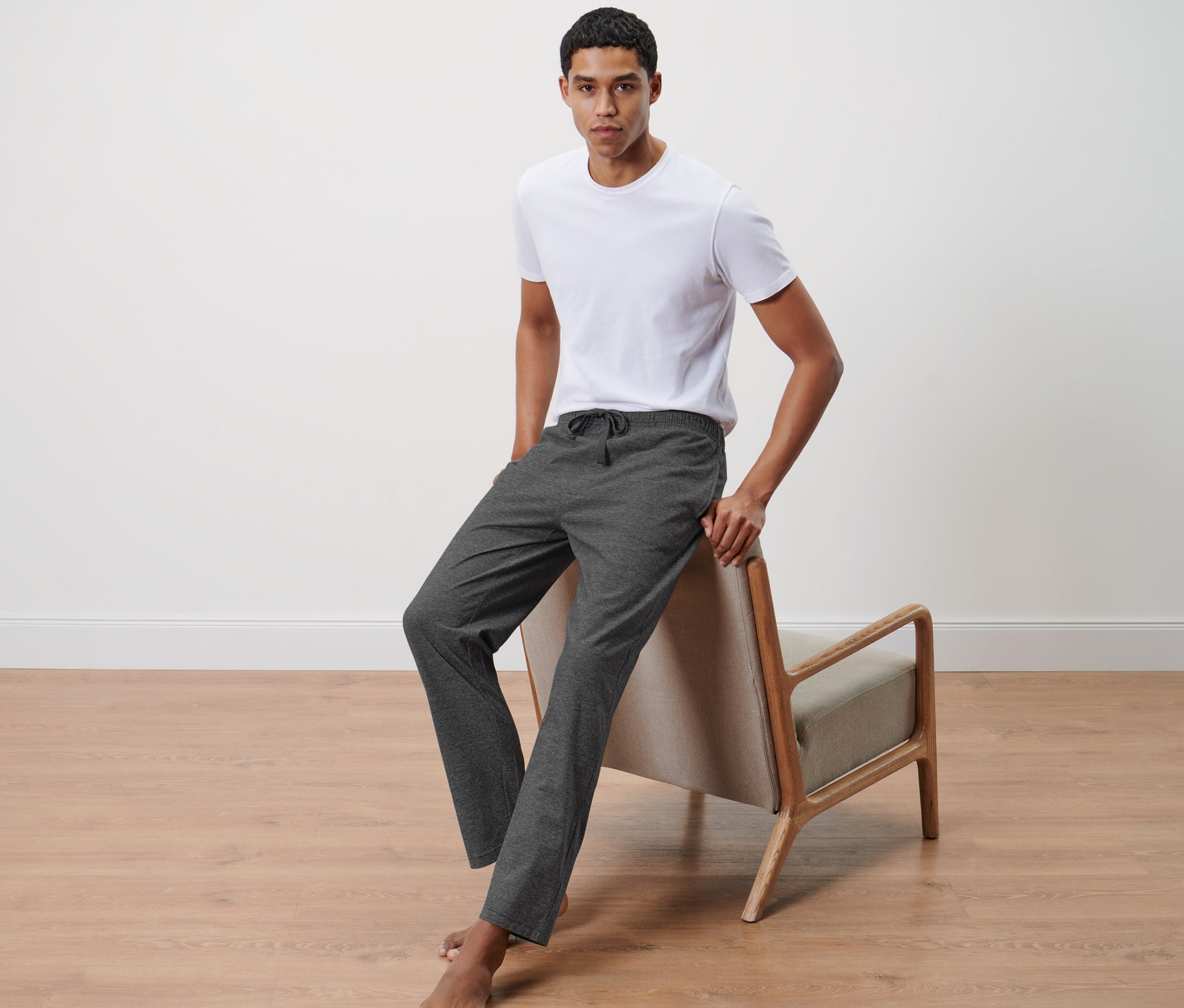 Muž opretý o stoličku má na sebe biele tričko a pohodlné nohavice.