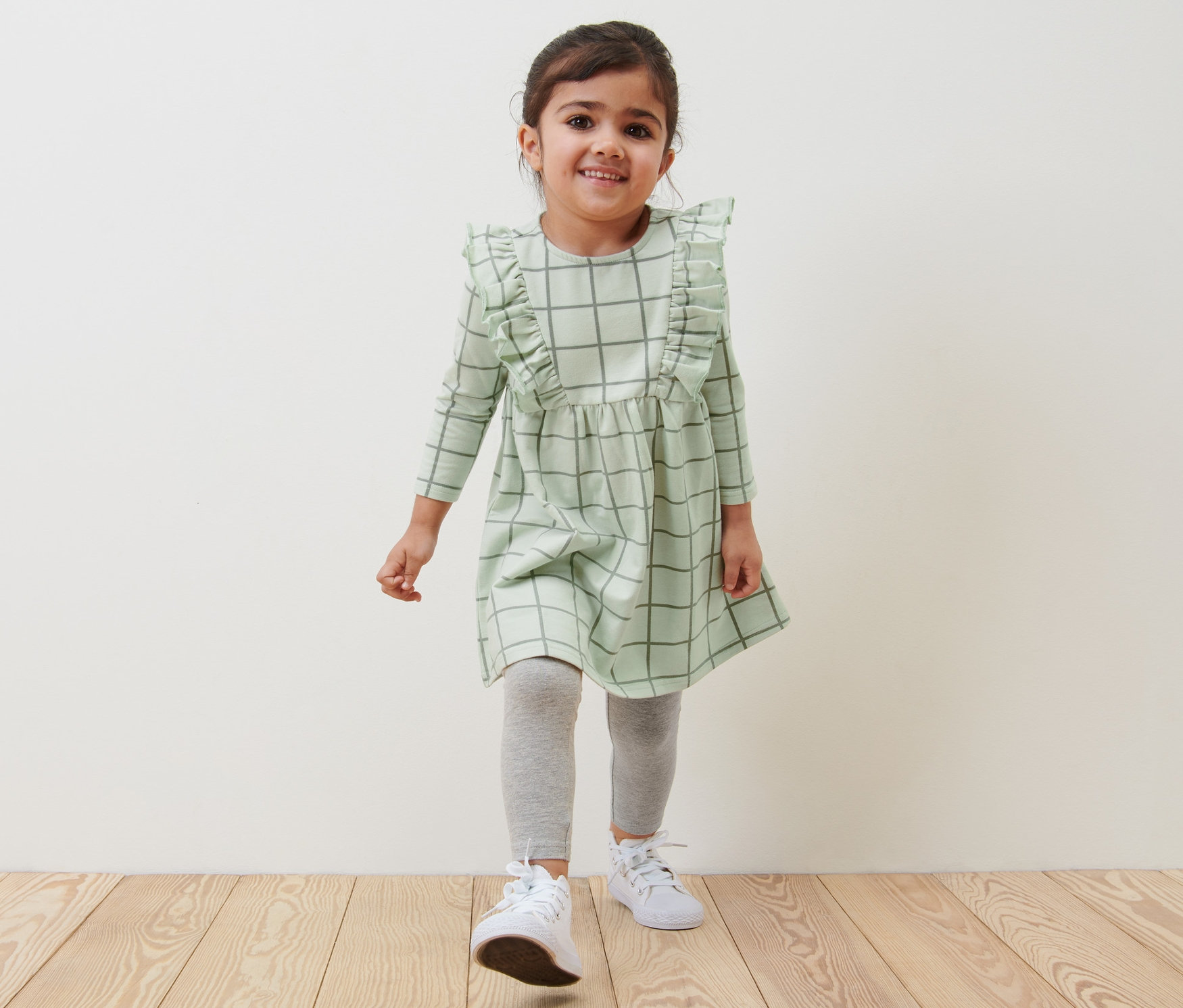 Usmievajúce sa dievčatko kráča po drevenej podlahe v detských teplákových šatách.