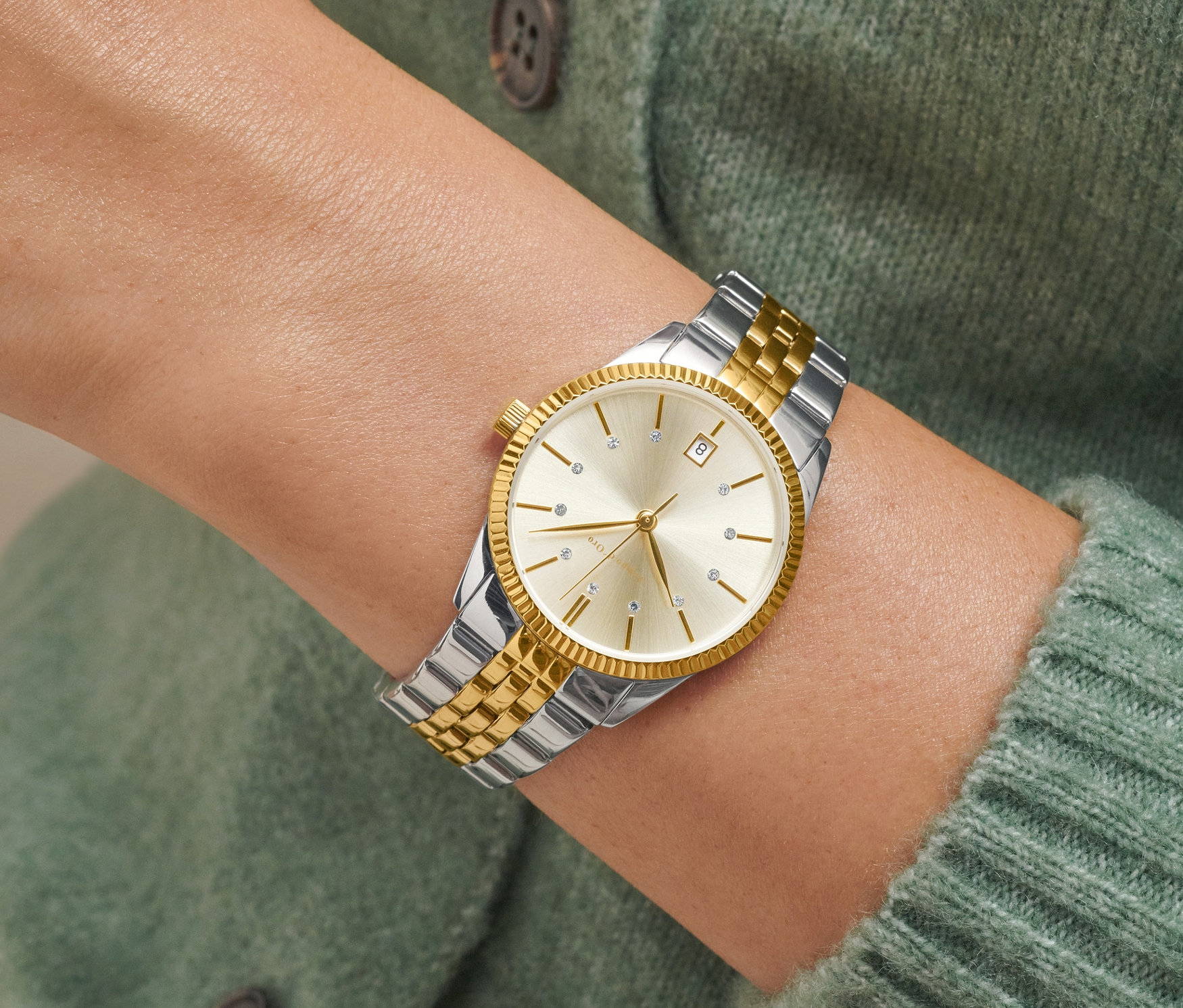 Detail dvojfarebných dámskych hodiniek s dátumom na zápästí ženy v zelenom svetri.