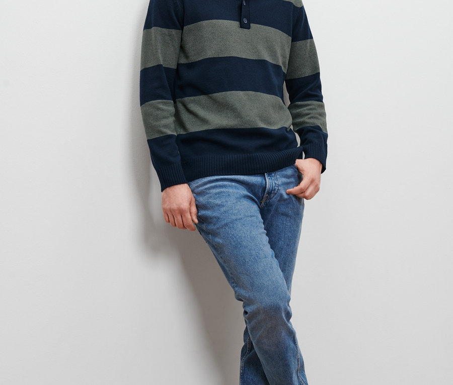 Muž opretý o stenu má na sebe pruhovaný pulóver a džínsy.