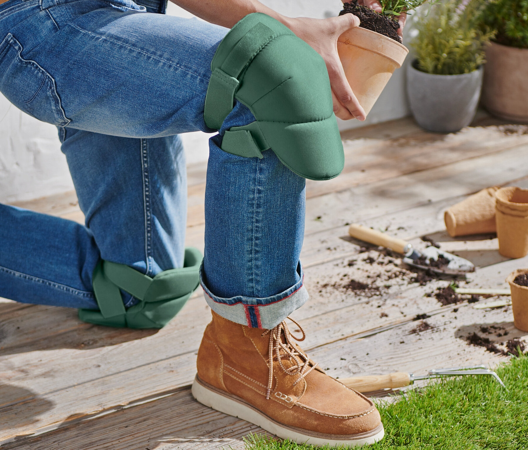 Osoba kľačí s nasadenými chráničmi kolien na záhradnícke práce a drží kvetináč s rastlinou.