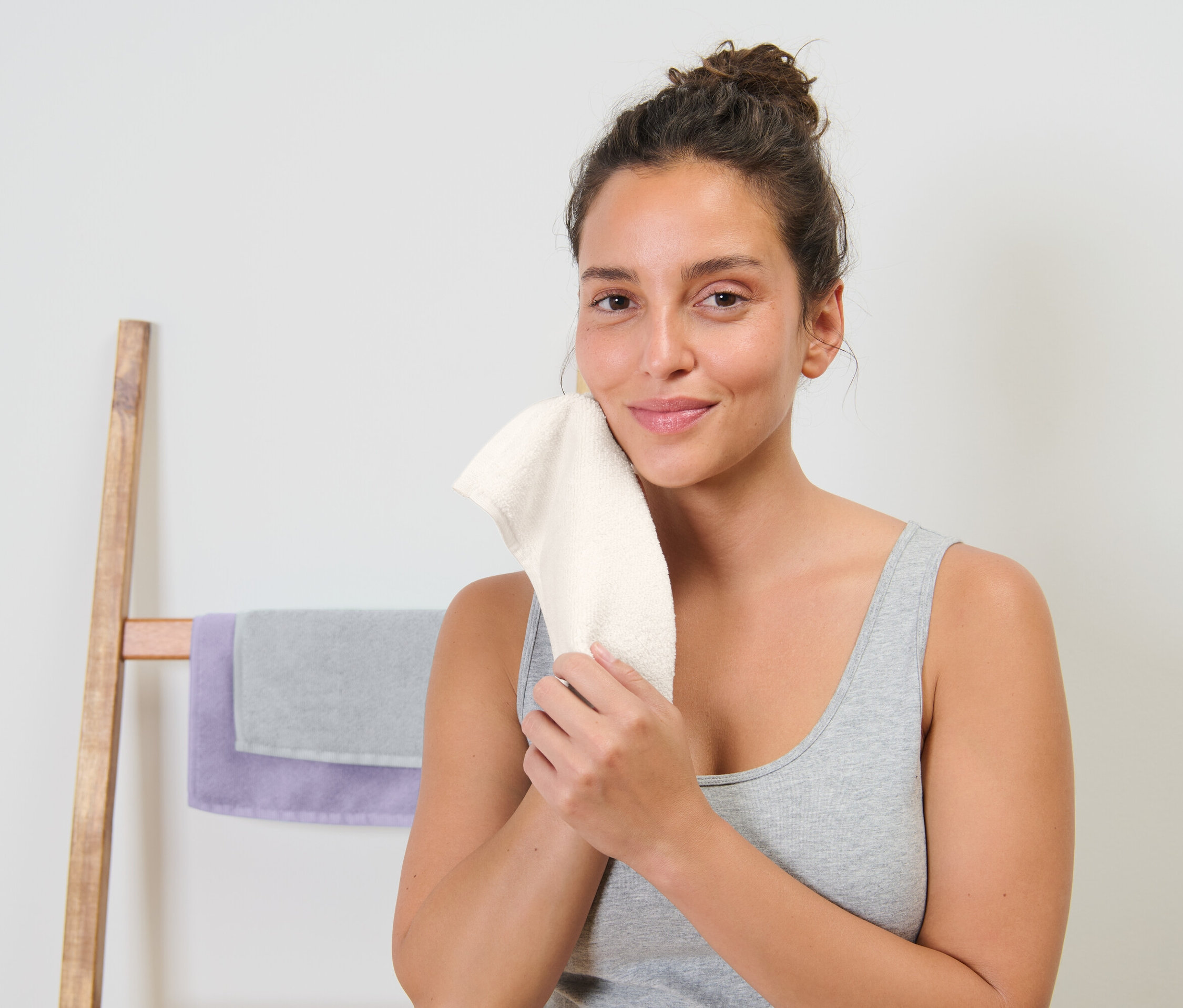 Žena si utiera tvár bielym uterákom. Za ňou je drevený vešiak na uteráky so sivým a fialovým uterákom.