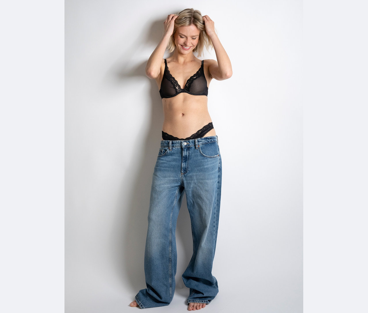 Žena sa usmieva a drží si ruky na hlave, má na sebe čiernu čipkovanú podprsenku a tangá a obrovské džínsy.