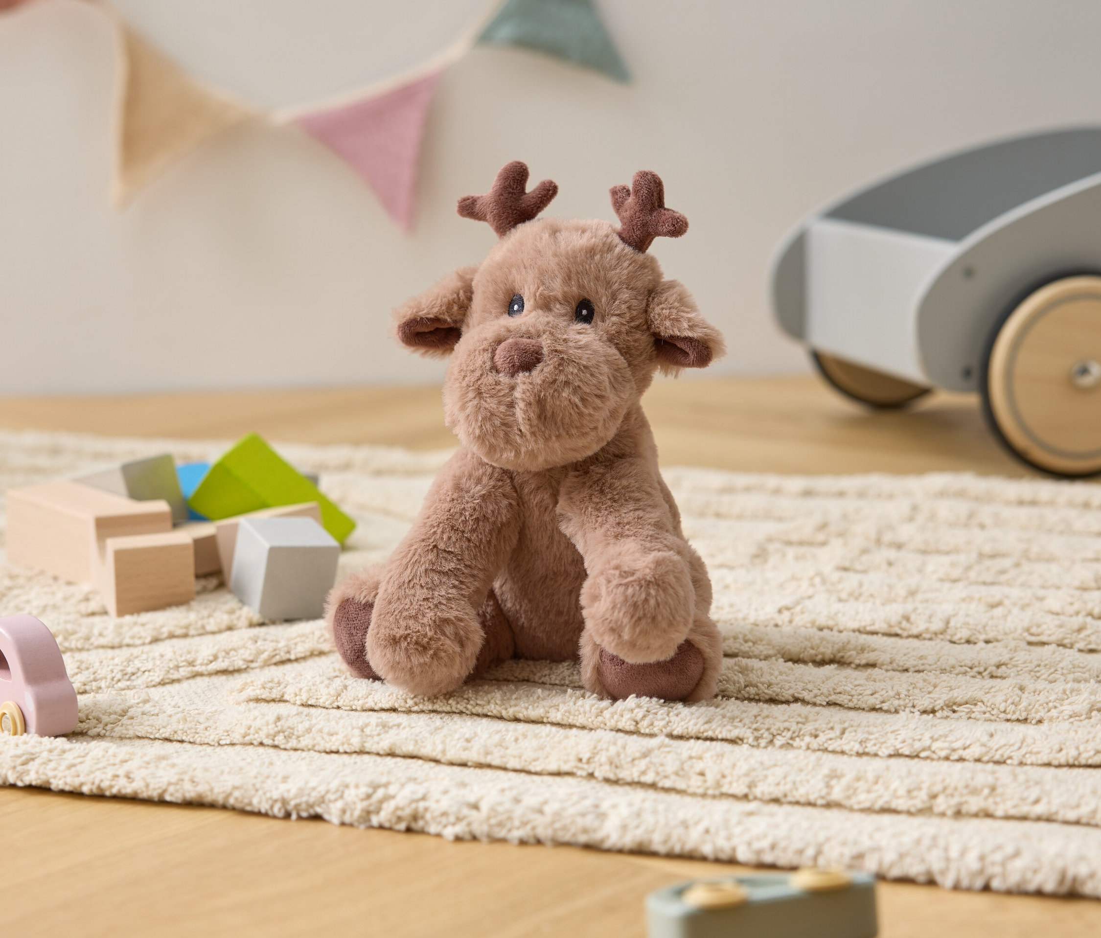 Plyšový los sedí na krémovom koberci medzi drevenými hračkami a sivým vozíkom.