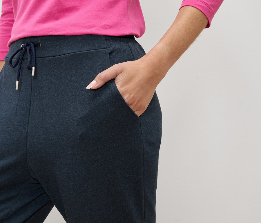 Detail osoby v tmavomodrých teplákoch so šnúrkou a vreckami, s rukou vo vrecku.