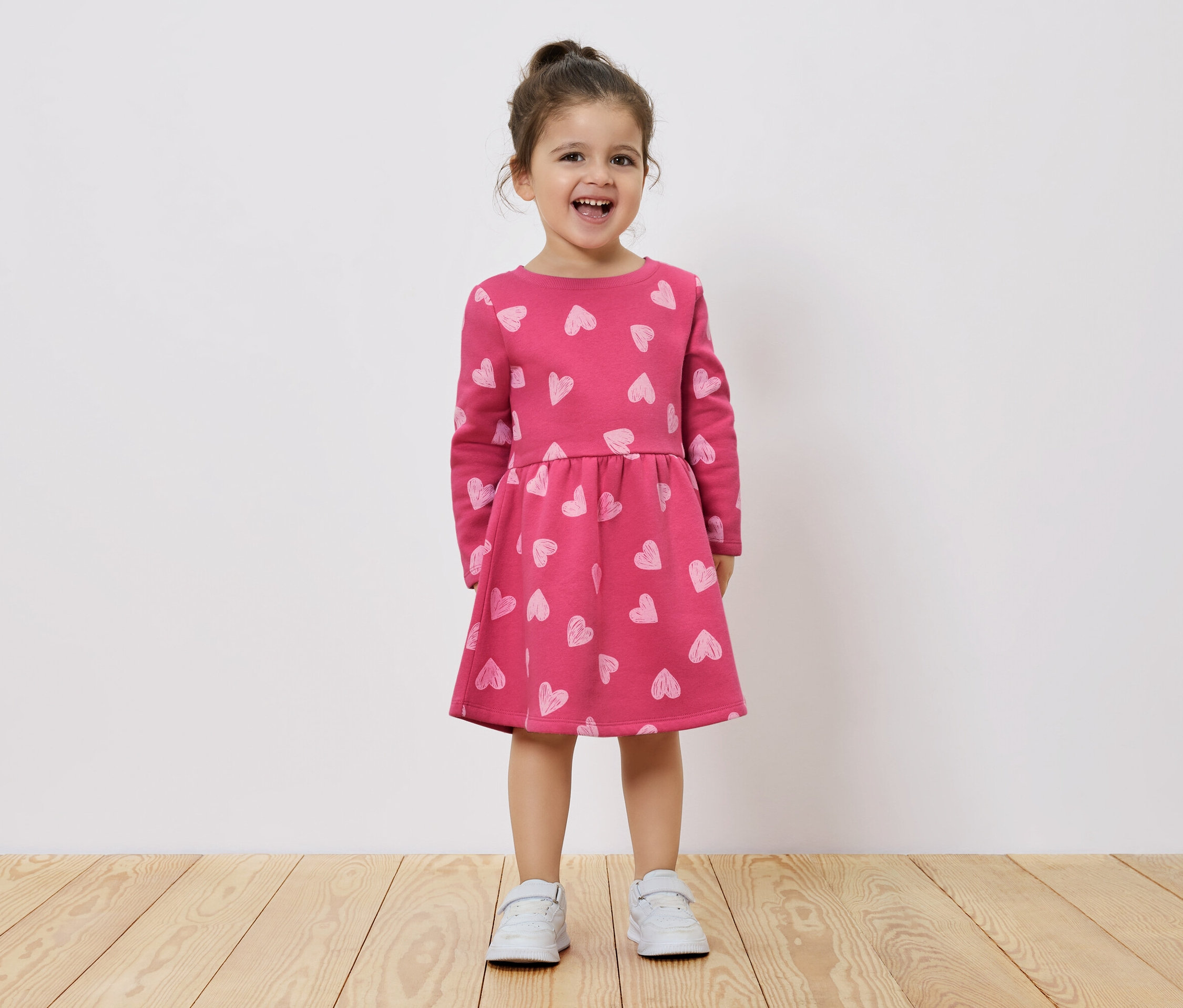 Usmievajúce sa dievčatko stojí na drevenej podlahe a má na sebe ružové šaty s dlhým rukávom a potlačou srdiečok.