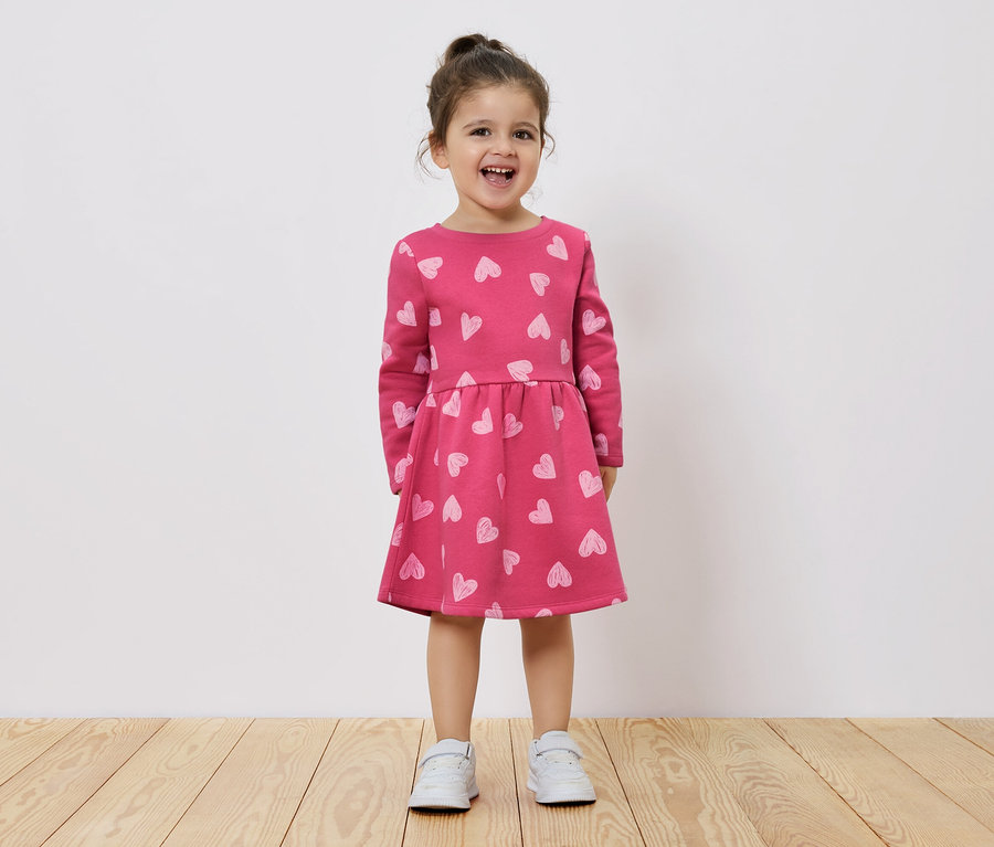 Usmievajúce sa dievčatko stojí na drevenej podlahe a má na sebe ružové šaty s dlhým rukávom a potlačou srdiečok.