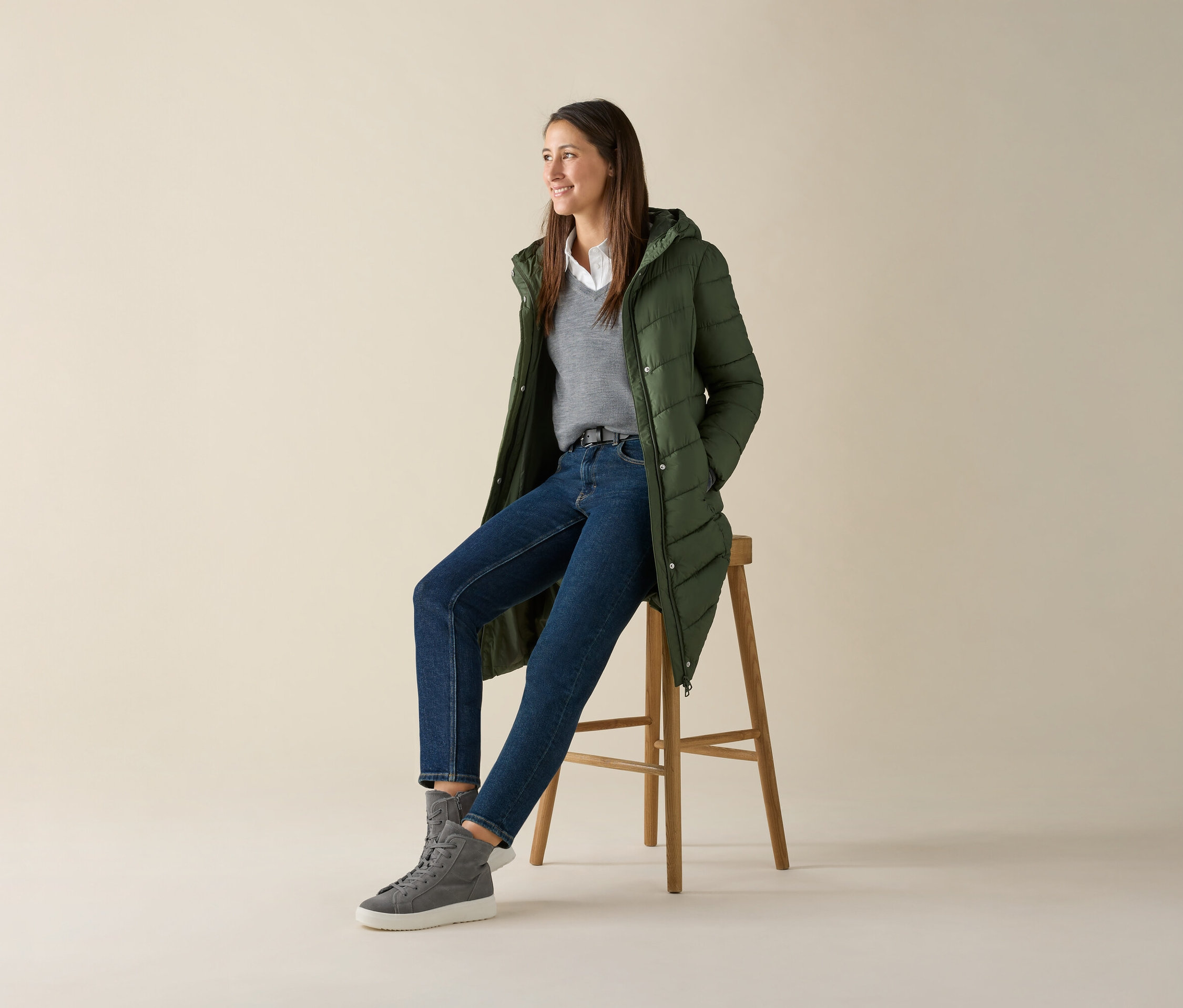 Žena sedí na stoličke a má na sebe sivý pulóver z jemného úpletu s merino vlnou, džínsy a zelenú bundu.