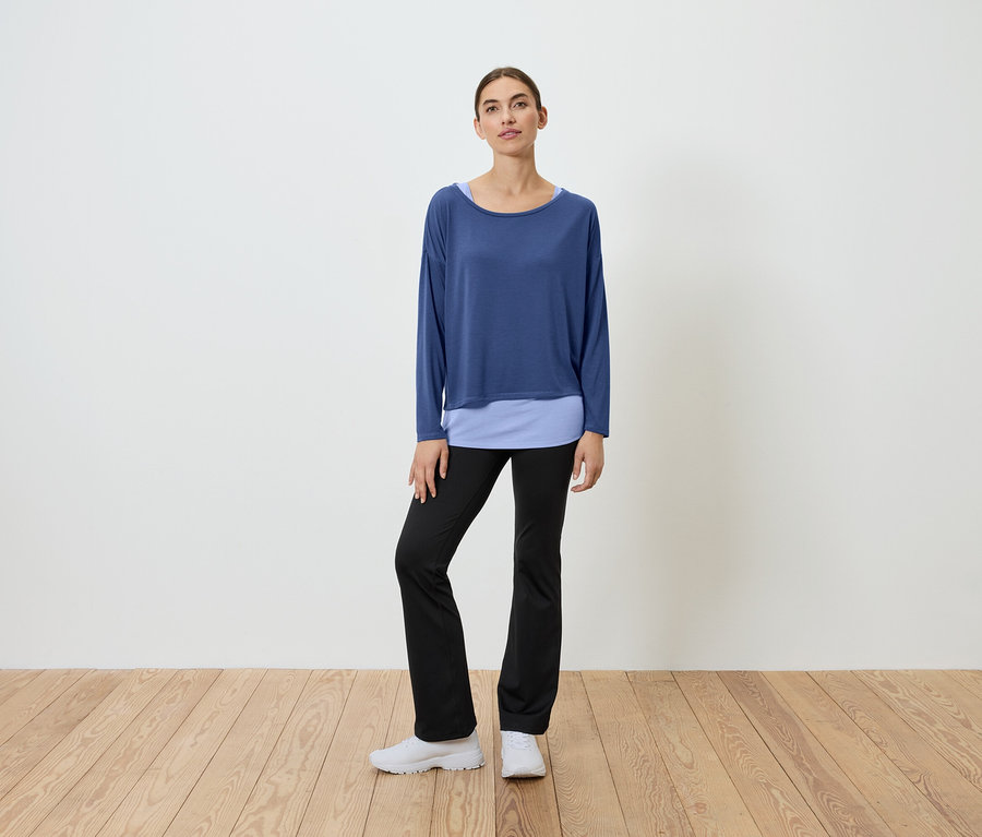 Žena stojí na drevenej podlahe a má na sebe modrý top a čierne nohavice.