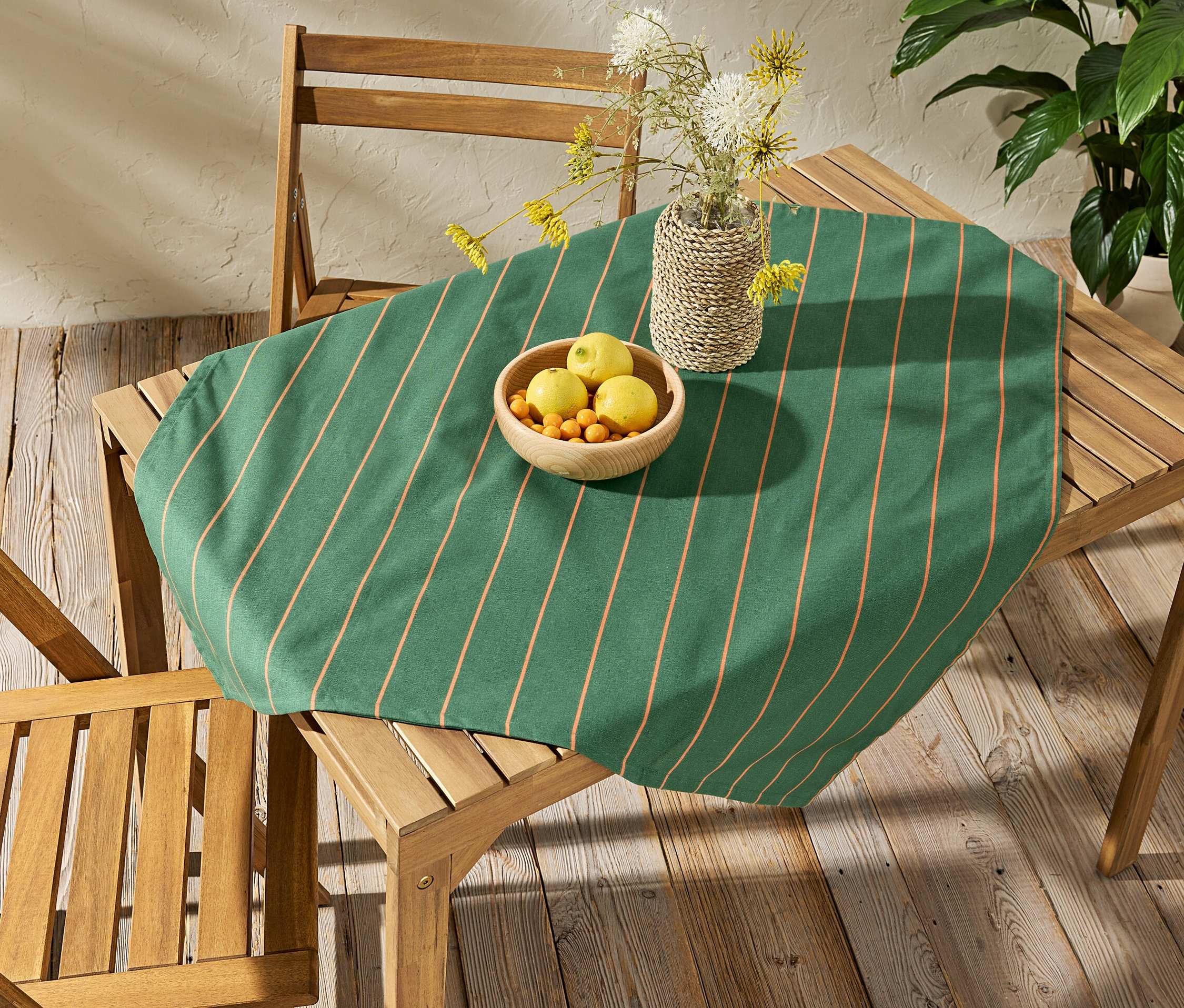 Na drevenom stole leží umývateľný obrus so vzorom prúžkov, cca 90 x 90 cm. Na obruse je miska s ovocím a váza s kvetmi.