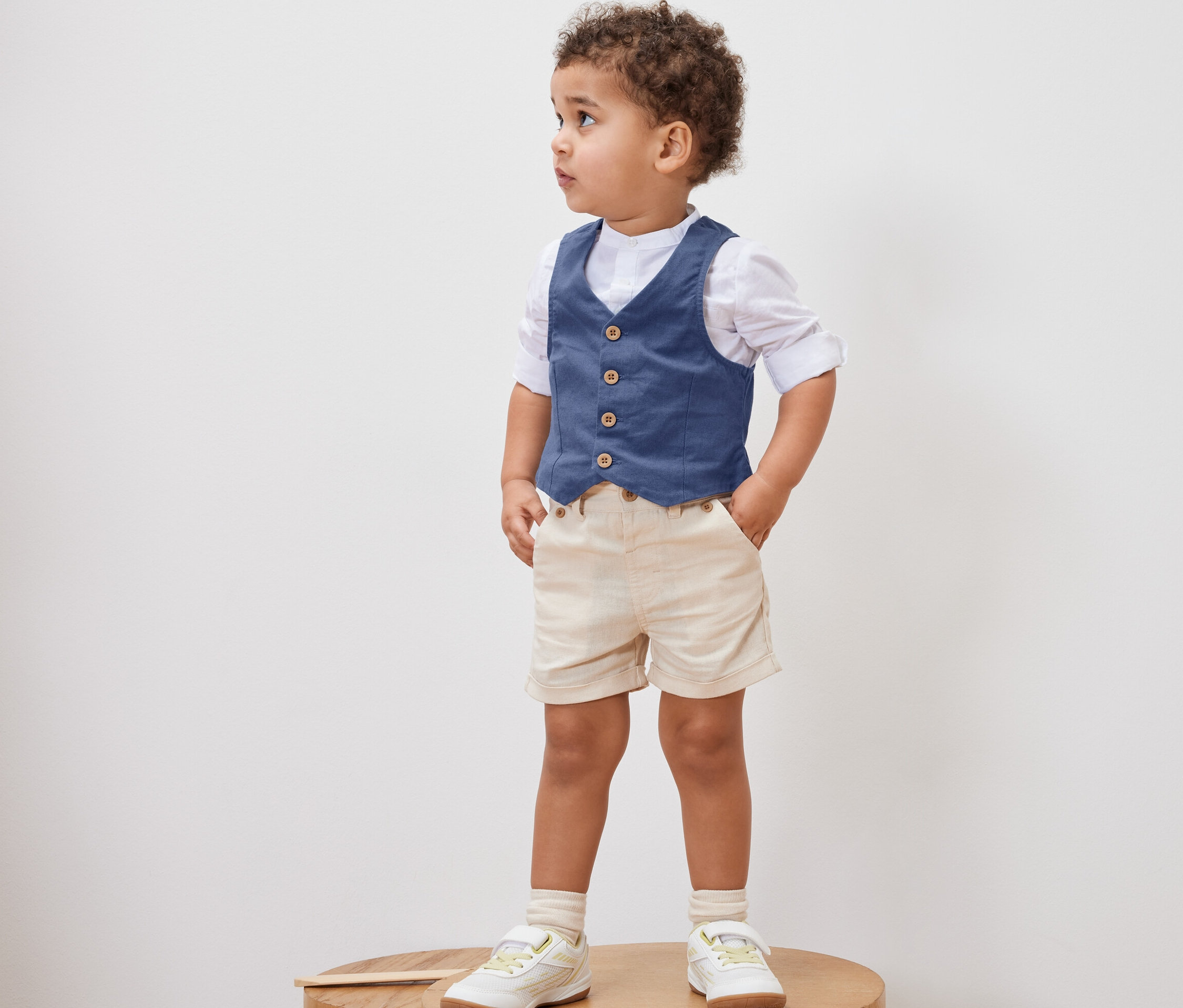 Chlapček stojí na drevenom podstavci a má na sebe Detskú vestu, košeľu, kraťasy a topánky.