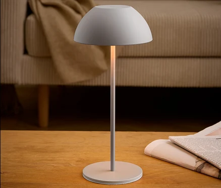 Sivá lampa stojí na drevenom stole vedľa novín. V pozadí je béžová pohovka.