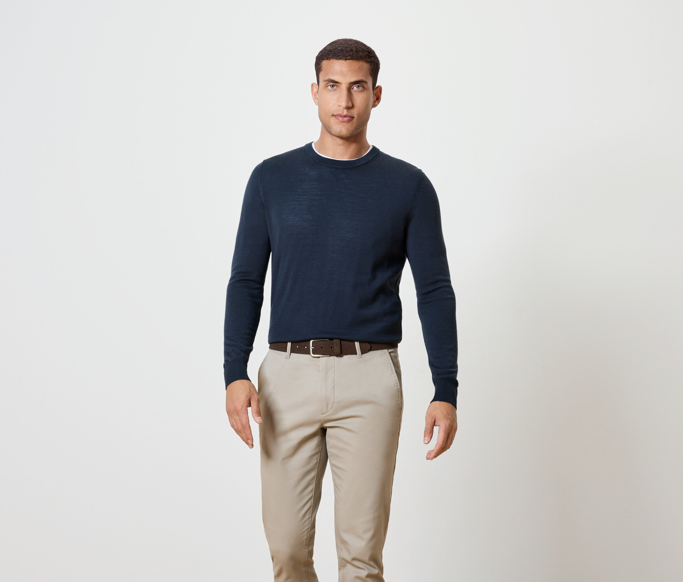 Muž stojí a má na sebe tmavomodrý sveter, hnedý opasok a khaki nohavice.