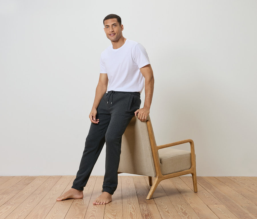 Muž opretý o stoličku, má na sebe biele tričko a sivé tepláky.