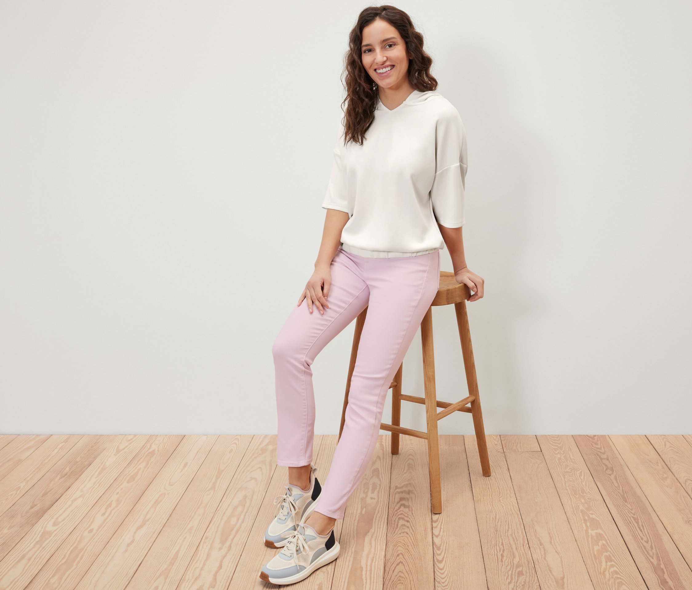 Žena sedí na stoličke a má na sebe krémovobielu mikinu s kapucňou z modalovej zmesi a ružové nohavice.