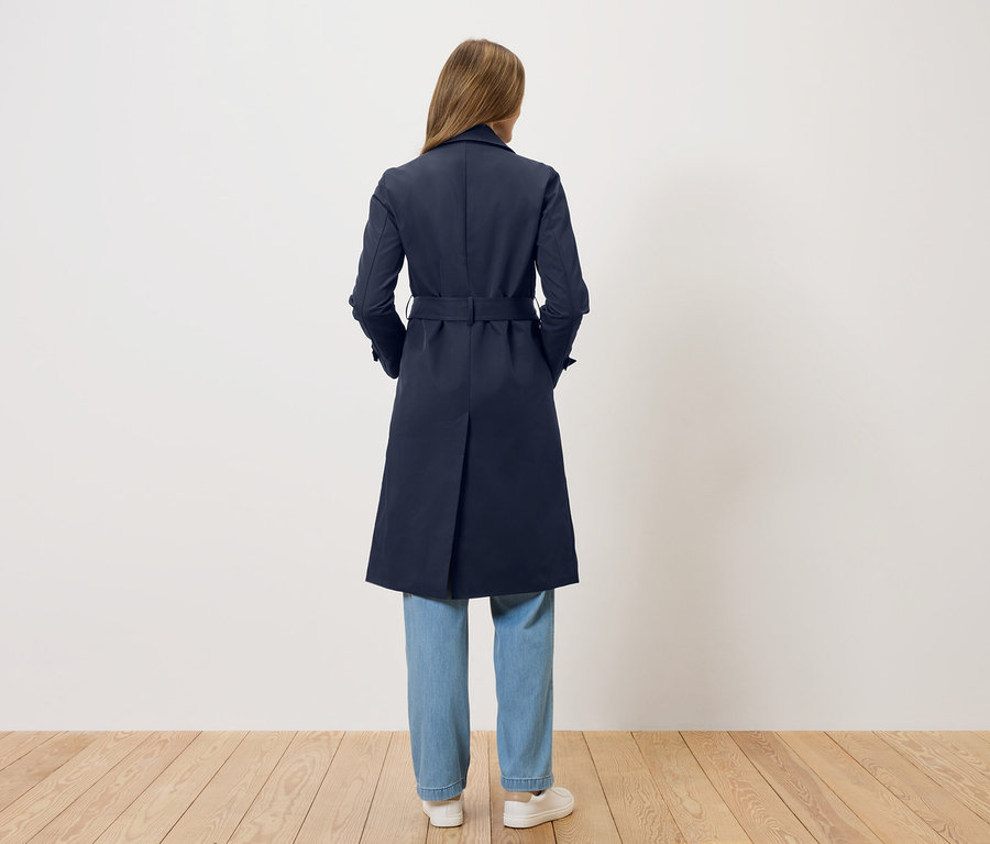 Žena stojí chrbtom k fotoaparátu a má na sebe tmavomodrý trenčkot, džínsy a biele topánky.