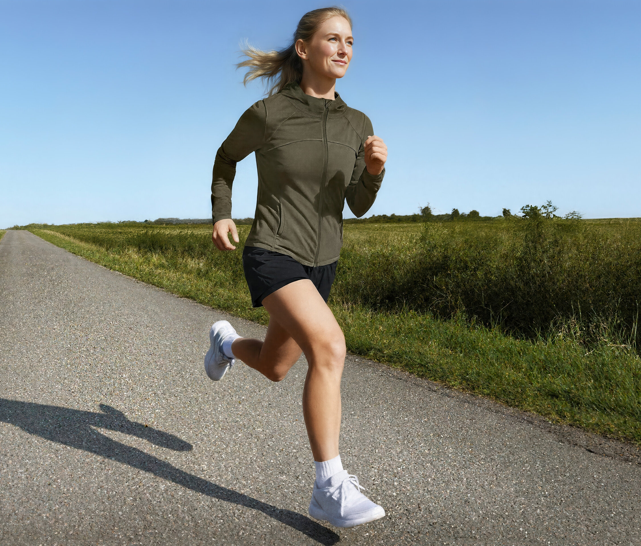 Žena beží v športovej bunde a 2-v-1 bežeckých šortkách po vidieckej ceste.