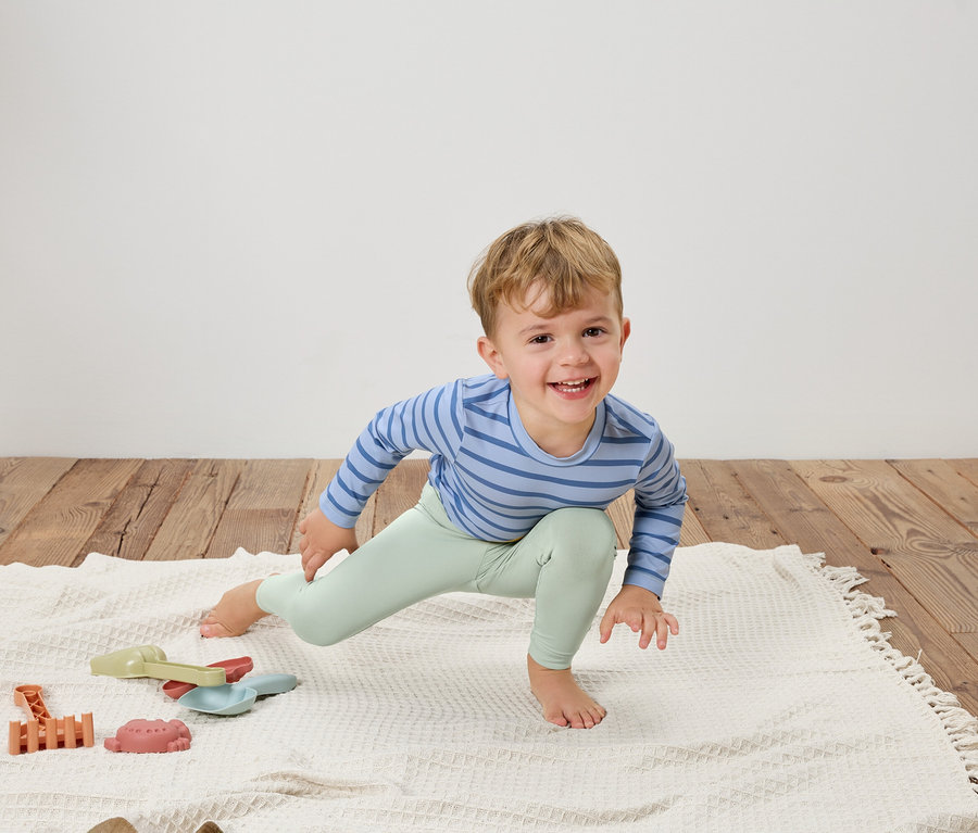 Usmievavý chlapec v modro pruhovanom tope a svetlozelených legínach sa naťahuje na bielej deke vedľa hračiek.