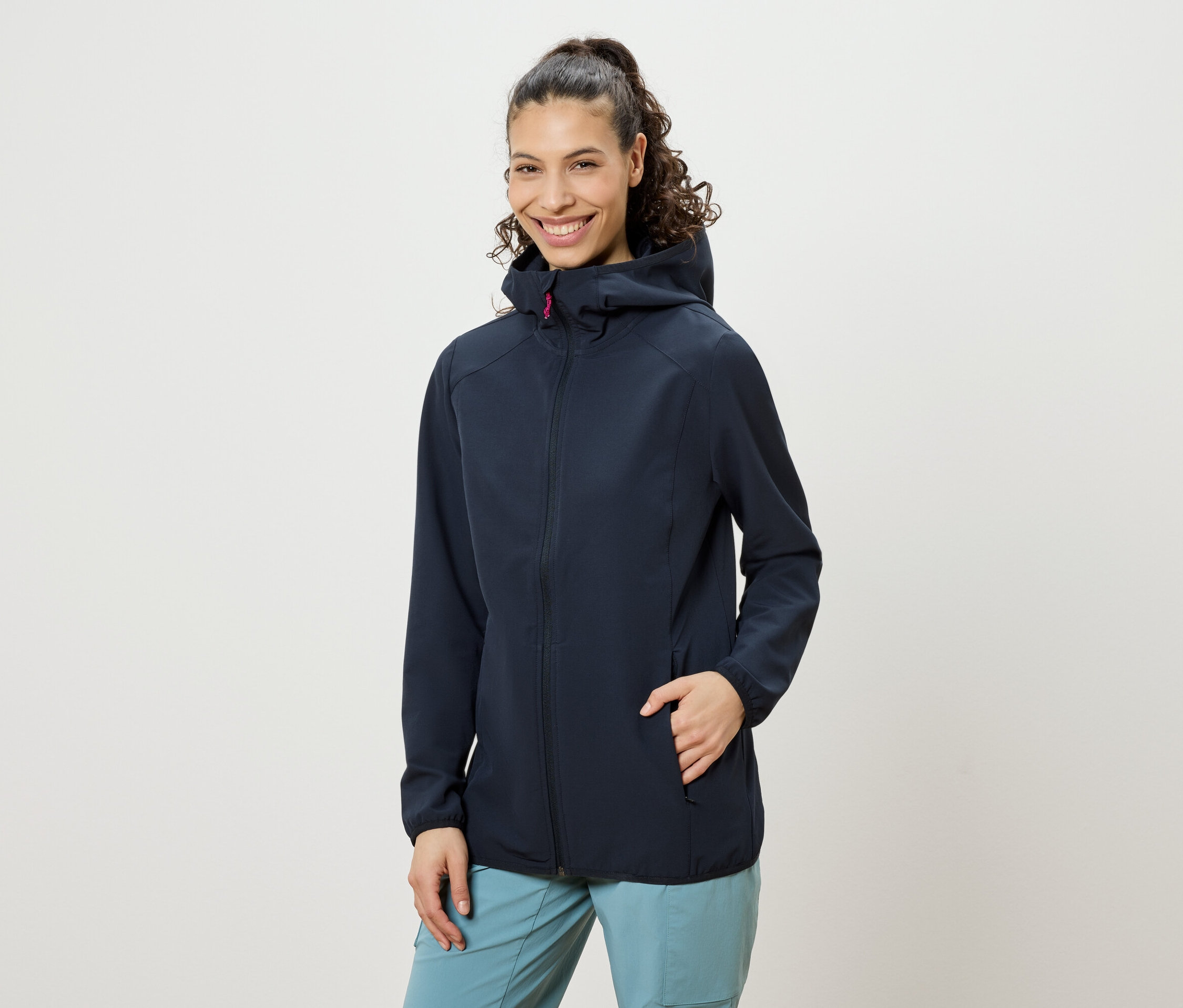 Žena s kučeravými vlasmi zopnutými do copu, oblečená v tmavomodrej bunde s kapucňou a modrých nohaviciach, usmieva sa, pričom má jednu ruku vo vrecku bundy.