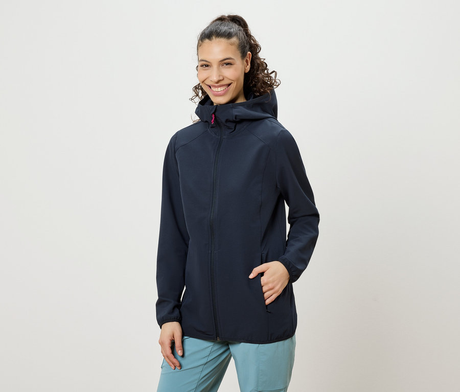 Žena s kučeravými vlasmi zopnutými do copu, oblečená v tmavomodrej bunde s kapucňou a modrých nohaviciach, usmieva sa, pričom má jednu ruku vo vrecku bundy.