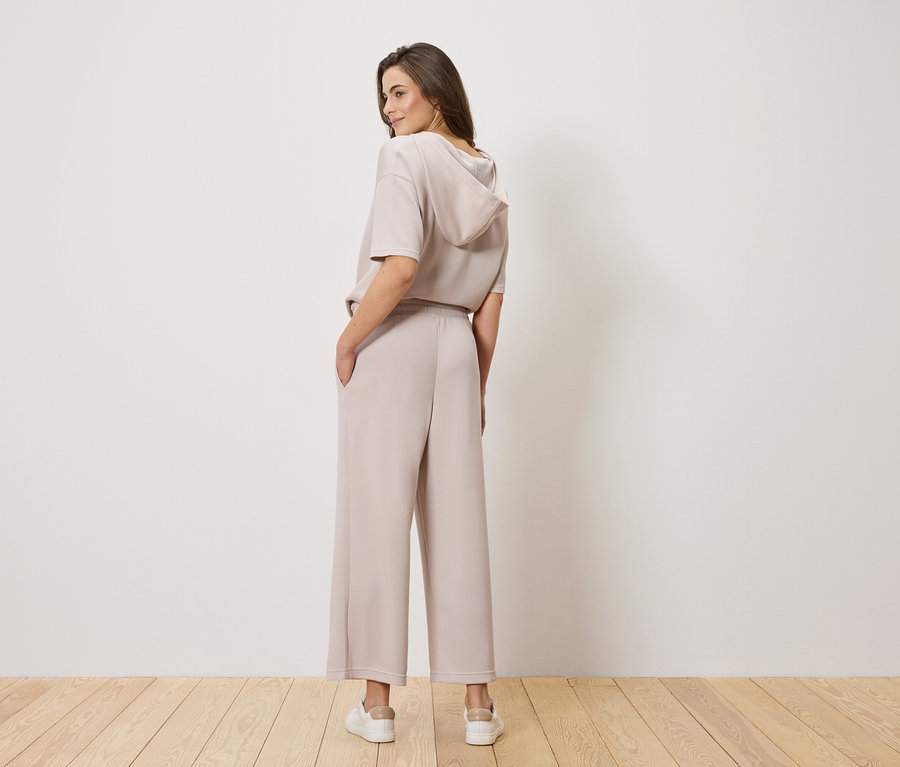 Žena s hnedými vlasmi, fotená zozadu, má na sebe svetlohnedý top s kapucňou a široké nohavice.