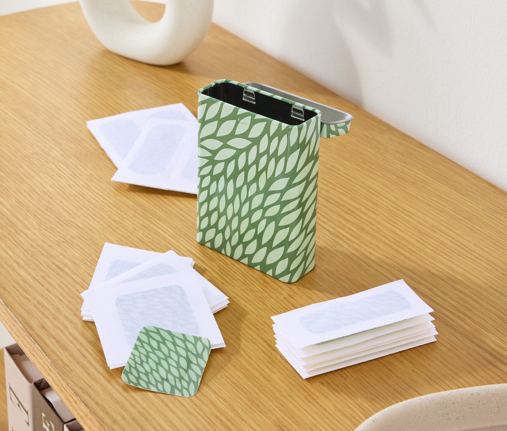 Na drevenom stole je plechovka so zeleným vzorom listov, prázdne voskové pásiky a váza s rastlinami.