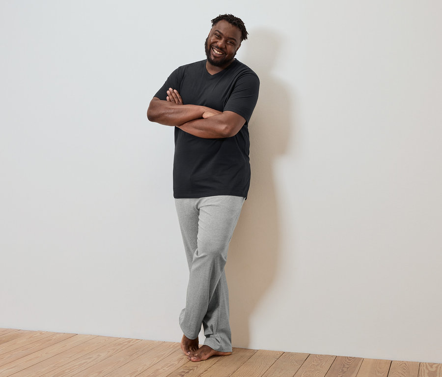 Tmavý muž so skríženými rukami sa opiera o stenu, má na sebe čierne tričko a sivé nohavice.