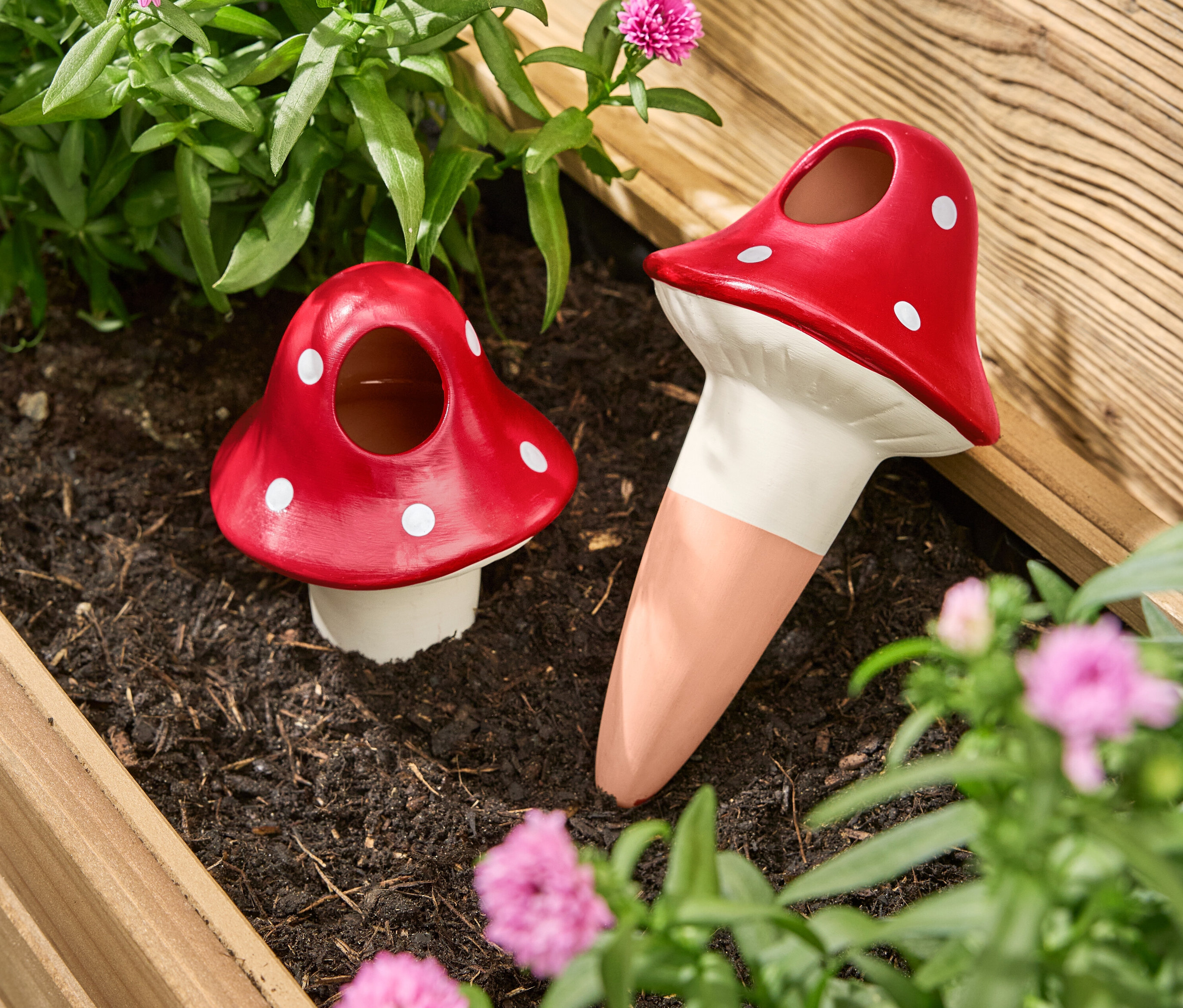 Dve červené zavlažovacie gule v tvare hríbov s bielymi bodkami stoja v kvetináči medzi kvetmi a rastlinami.