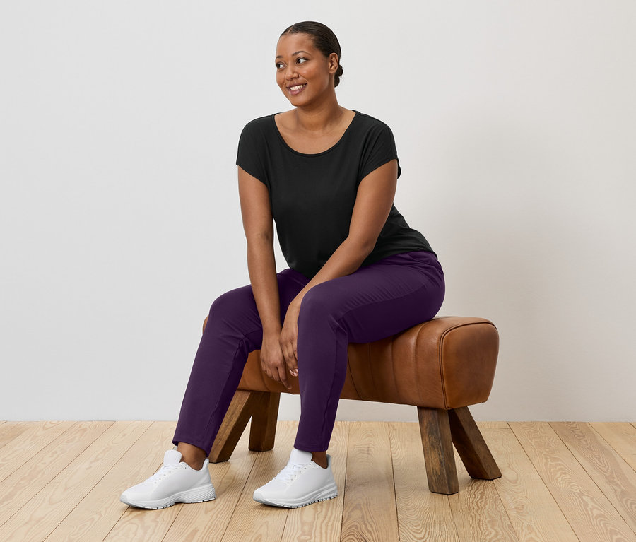 Žena s tmavými vlasmi sedí na gymnastickom koni a má na sebe čierne tričko, fialové nohavice a biele tenisky.