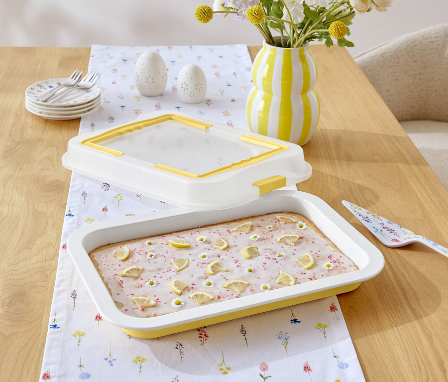 Na drevenom stole leží koláč s citrónovou polevou a margarétkami, žlto-biely pruhovaný džbán, lopatka na tortu s kvetinovým vzorom a biely obrus s kvetinovým vzorom.