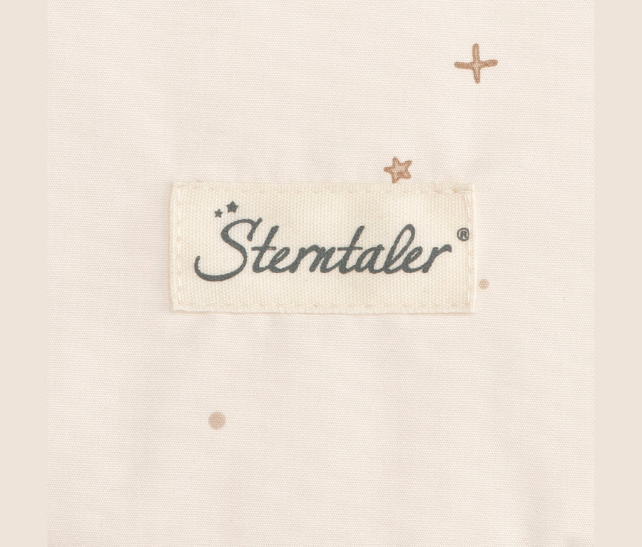 Detail svetlobéžovej látky s bielym našitým štítkom s tmavosivým logom 'Sterntaler'. Na látke sú roztrúsené malé svetlohnedé vzory krížikov, hviezdičiek a bodiek.