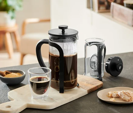 Čierna kanvica french press 800 ml, 6 šálok, naplnená kávou, stojí na mramorovej doske vedľa pohára kávy, misky s cukrom a sušienok. Vedľa je rozložená ďalšia kanvica french press.