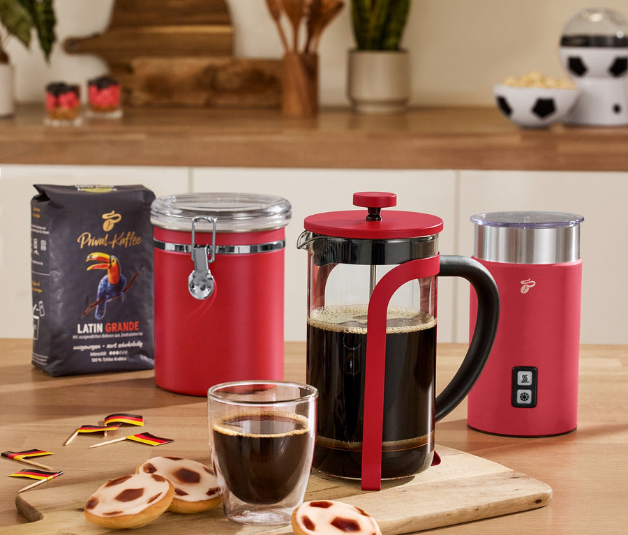 Na stole stojí červená dóza na zachovanie arómy, červený elektrický napeňovač mlieka, červená kanvica french press, 2 dvojstenné poháre na caffè crema a balenie kávy.