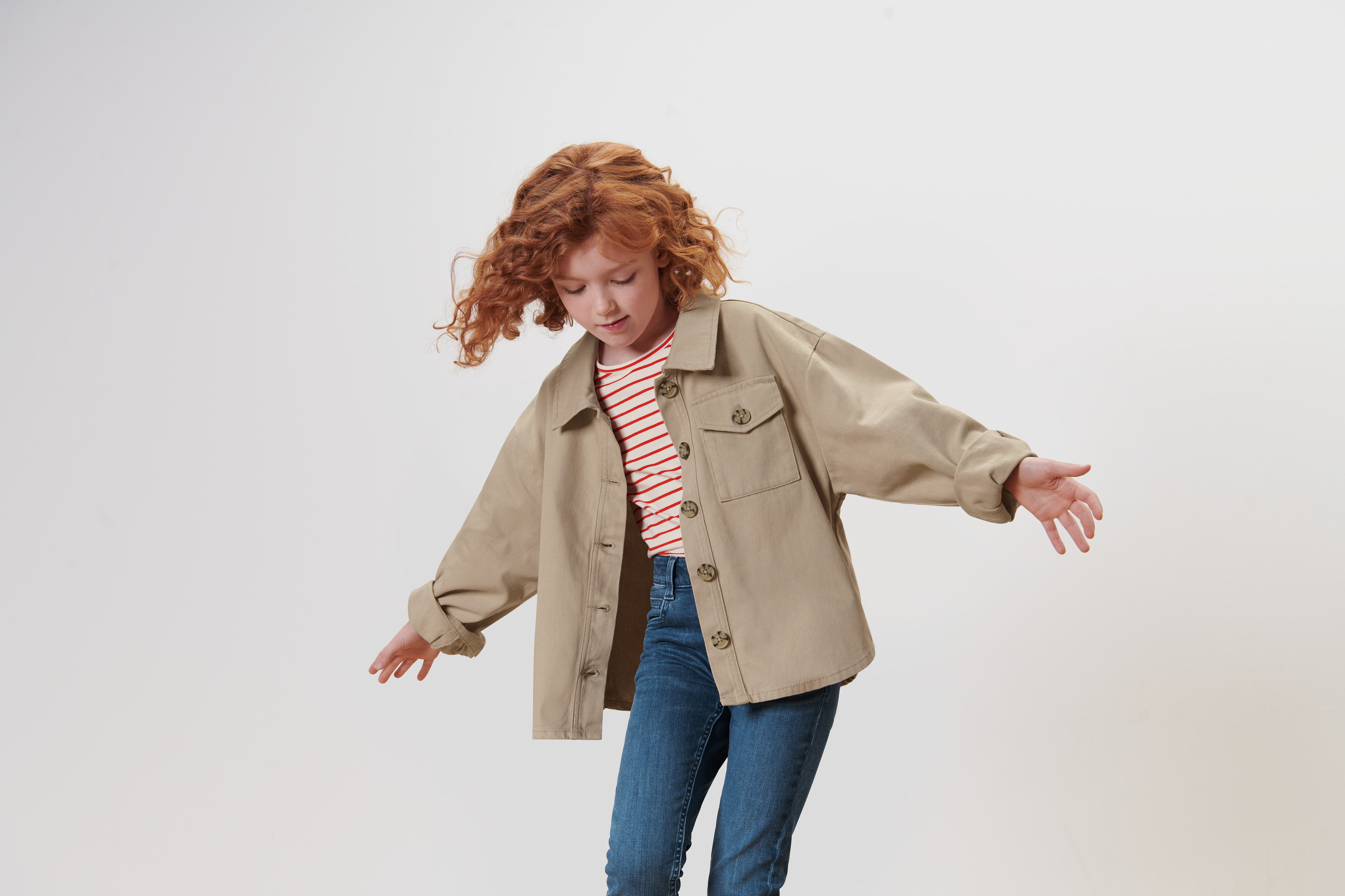 Dievča s kučeravými ryšavými vlasmi máva rukami a má na sebe detskú džínsovú bundu.