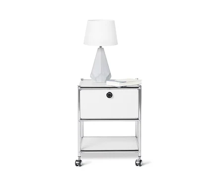 Na bielom pozadí je biely kovový odkladací stolík »CN3« so zásuvkou, na ktorom stojí lampa a leží kniha.