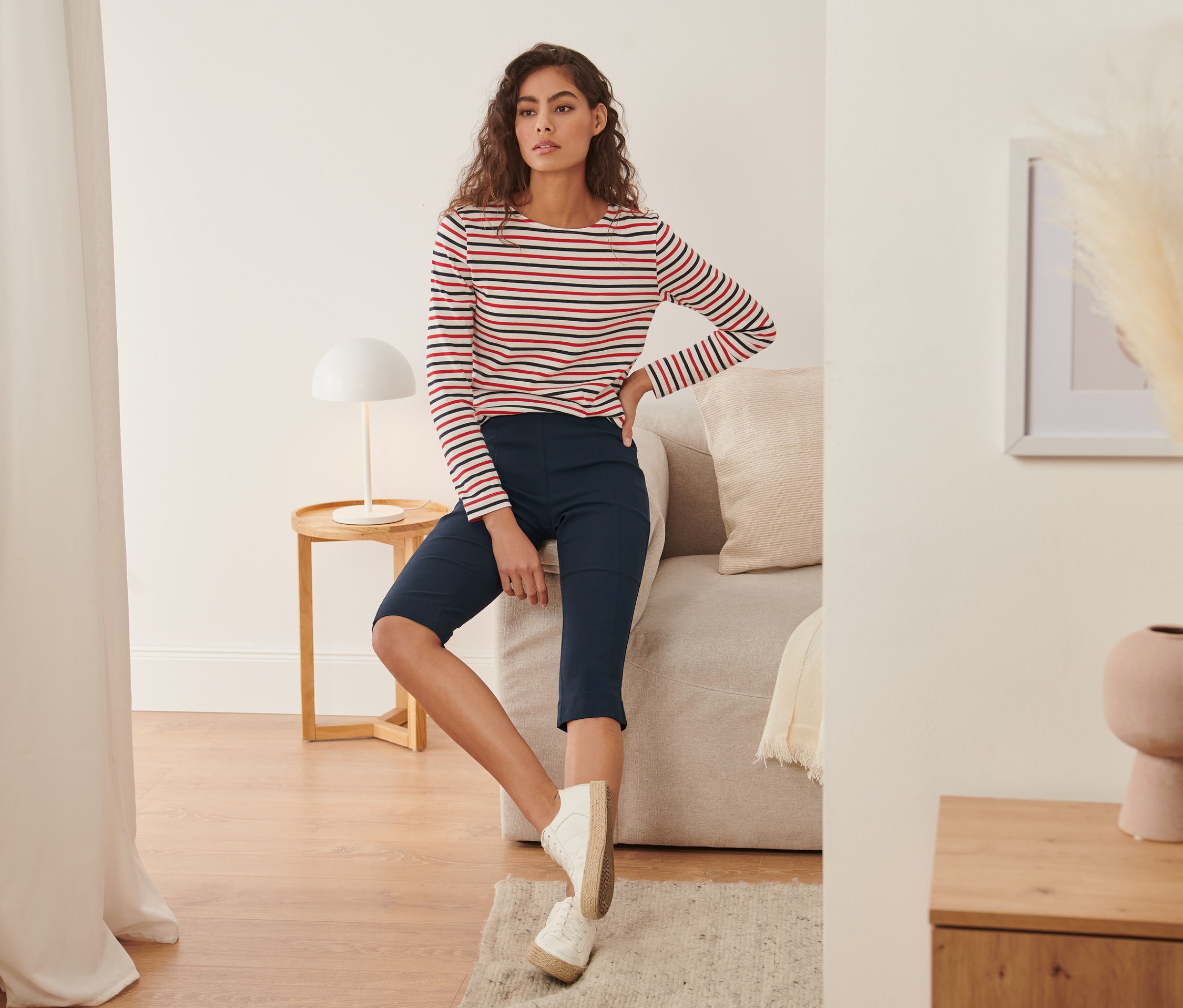 Žena sedí na pohovke a má na sebe trojštvrťové elastické nohavice a pruhované tričko.