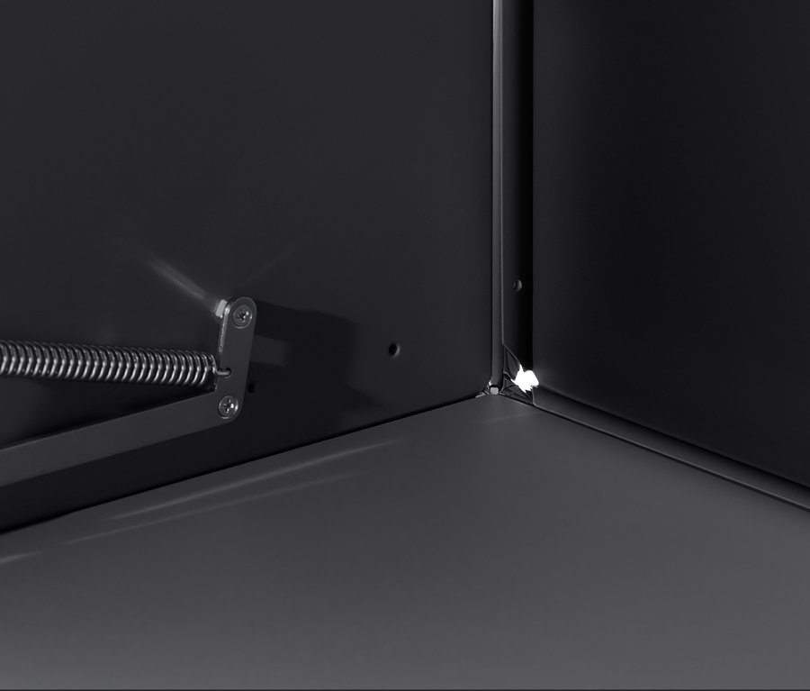 Detail vnútra čiernej skrinky »CN3« s výklopnými dvierkami, zameraný na roh a mechanizmus otvárania.