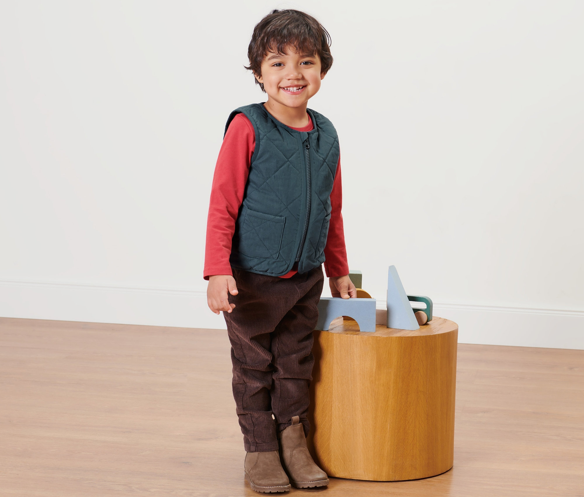 Chlapec stojí vedľa dreveného stolíka a má na sebe detskú obojstrannú prešívanú vestu.