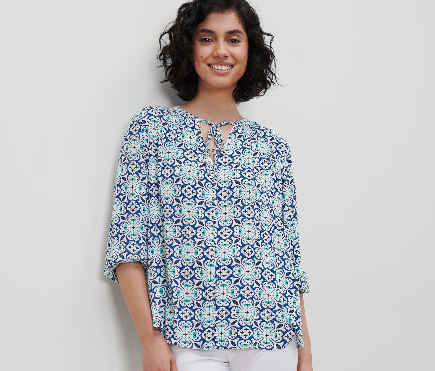 Žena má na sebe tunikovú blúzku s celoplošnou potlačou a biele nohavice.