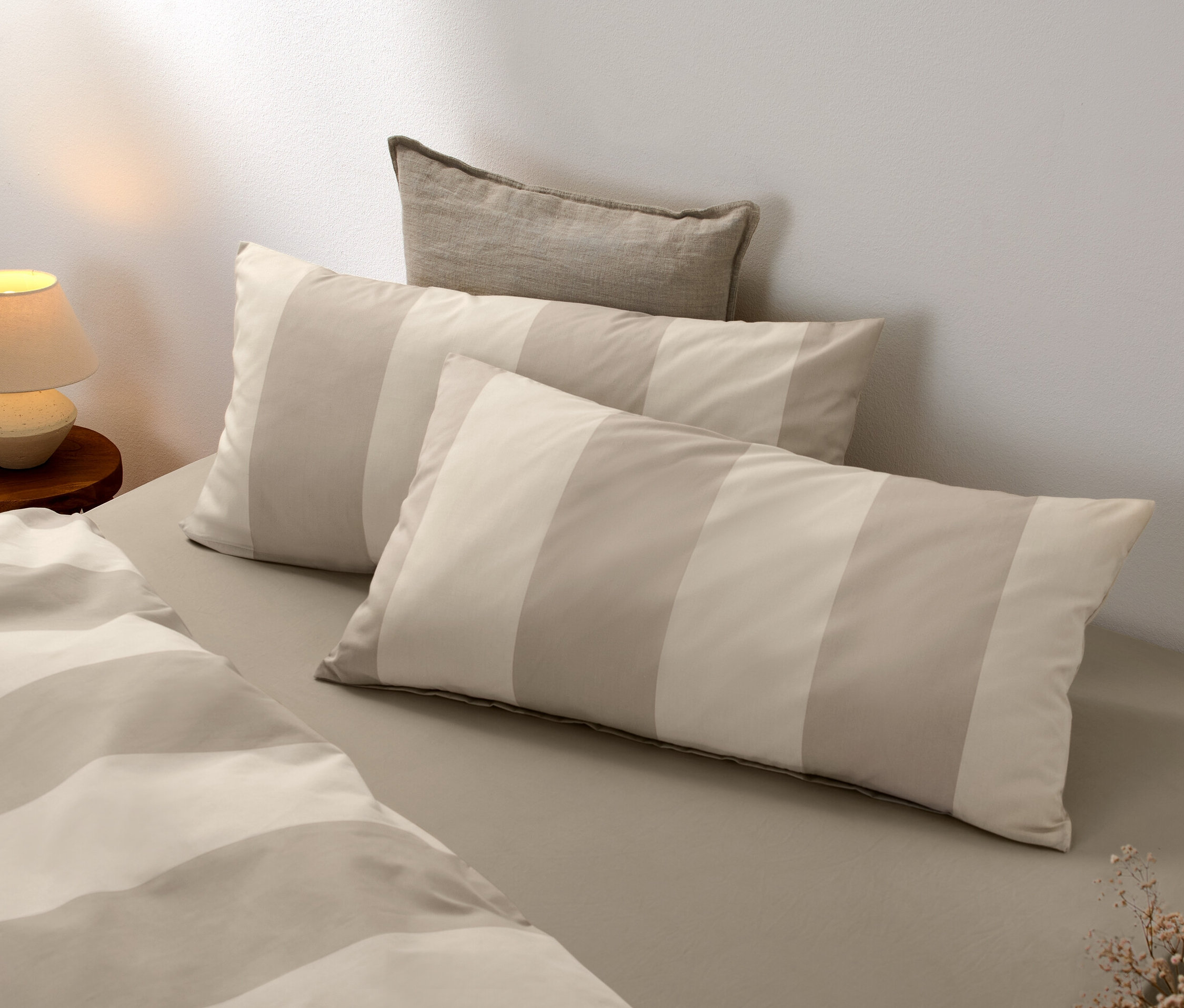 Dve pruhované béžové a krémové obliečky na vankúše z jemného flanelu ležia na posteli vedľa lampy.