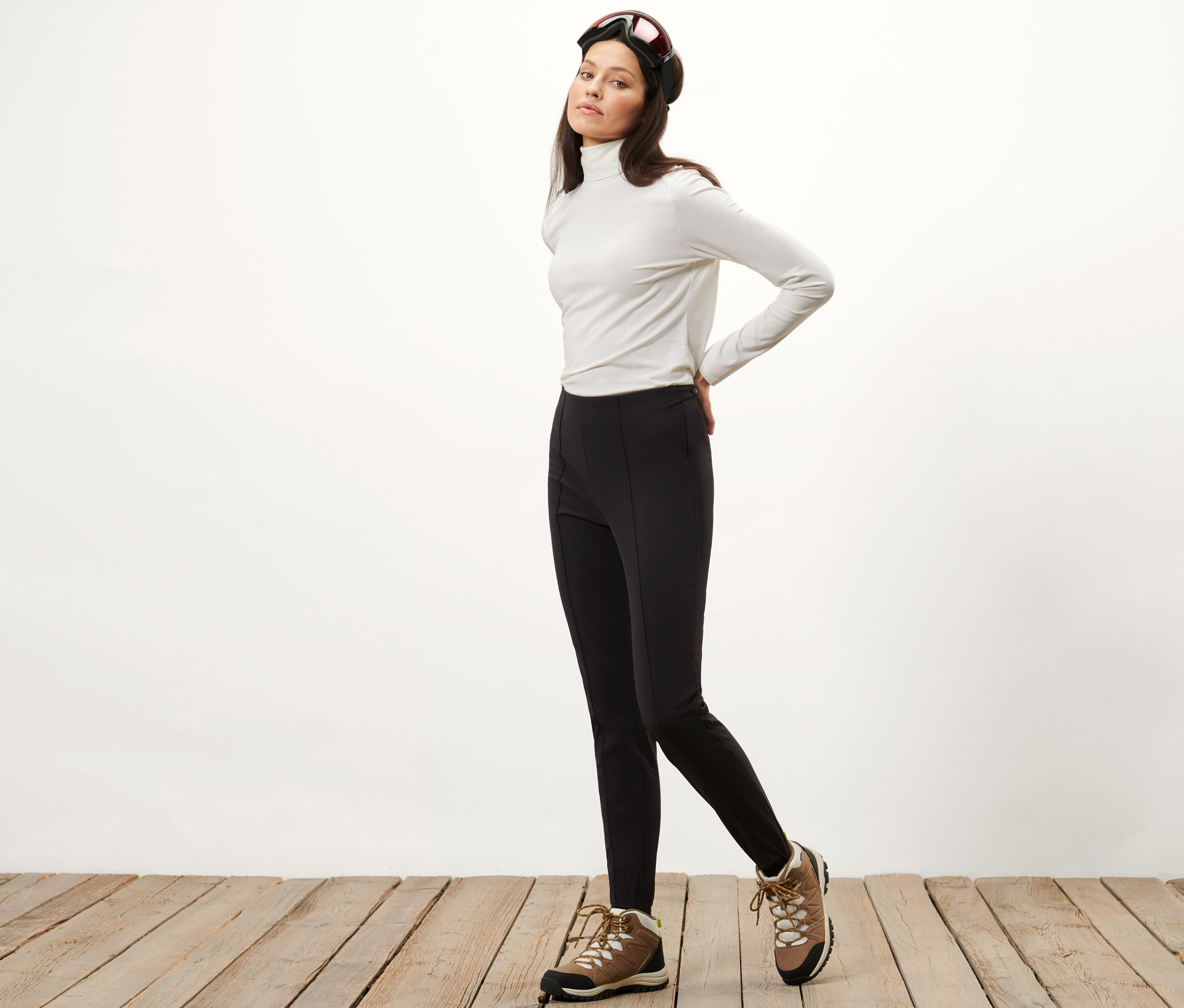 Žena pózuje v softshellových lyžiarskych priliehavých nohaviciach, bielom roláku a lyžiarskych okuliaroch.