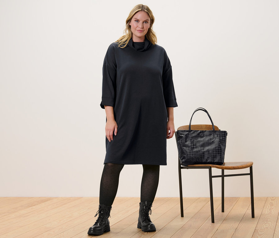 Žena stojí a má na sebe teplákové šaty z modalovej zmesi a pletené pančuchové nohavice so štruktúrou. Vedľa nej stojí stolička a na nej je kabelka.